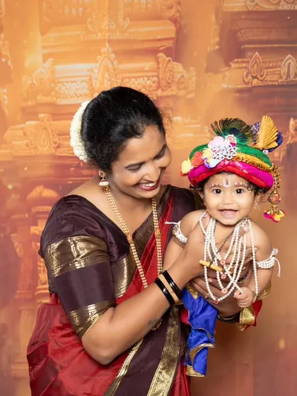 A mother dressed in a traditional saree holds her baby, who is playfully dressed as Lord Krishna for a themed photoshoot.