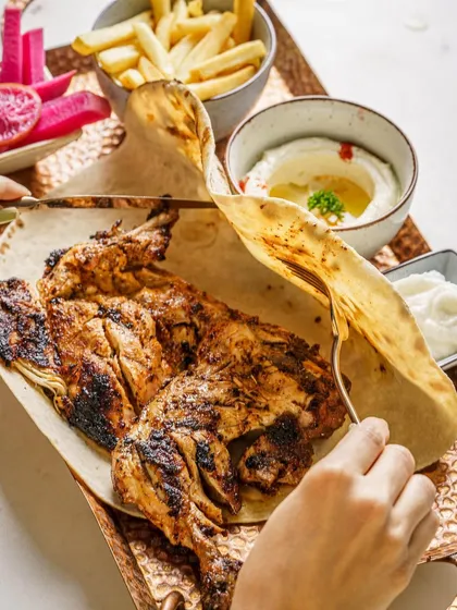 A whole grilled chicken served with hummus and pita. This shot captures a generous, shareable meal. The char marks on the chicken make it look flavorful and perfectly cooked.
