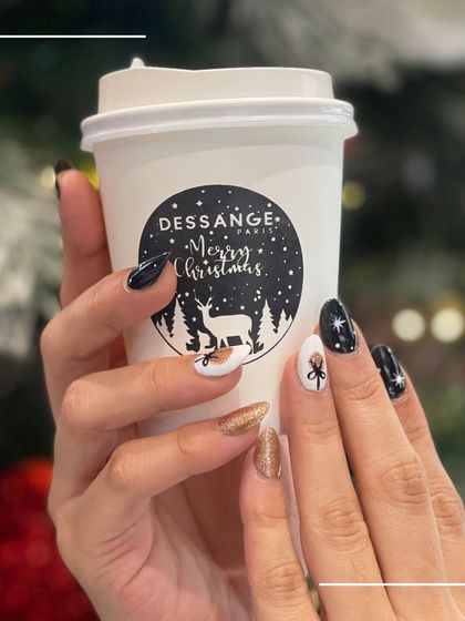 Holding a festive coffee, this client shows off her Christmas nail art, featuring black, white, and gold glitter with delicate bow and star designs.