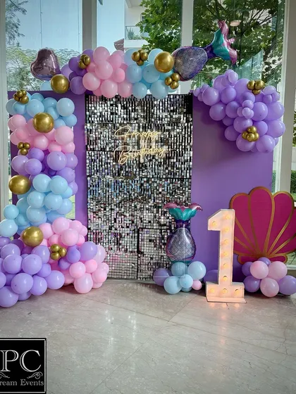 A glamorous mermaid-themed first birthday backdrop. This setup features a silver sequin wall, a 'Happy Birthday' neon sign, a light-up number '1', and a beautiful balloon arrangement in pastel purple, pink, and blue with gold accents.