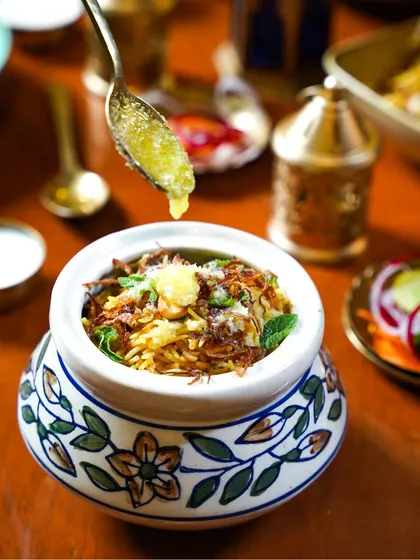 A rich and aromatic biryani in a traditional handi, with a spoonful of ghee being drizzled on top. The action and the warm lighting create a mouth-watering image for an Eid celebration.