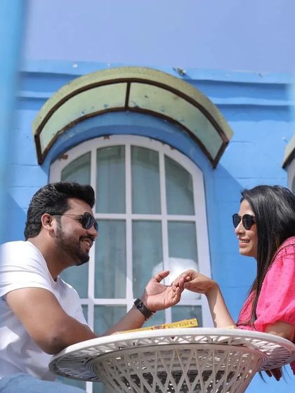 A casual and happy pre-wedding moment at an outdoor cafe setting. The bright blue background and the couple's genuine smiles make this a fresh and joyful photo.