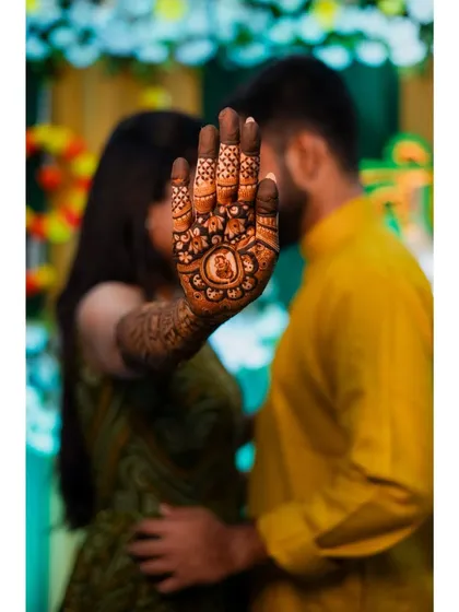 A romantic and creative mehendi photo, with the bride's henna-covered hand in the foreground and the couple sharing a quiet moment in the background.