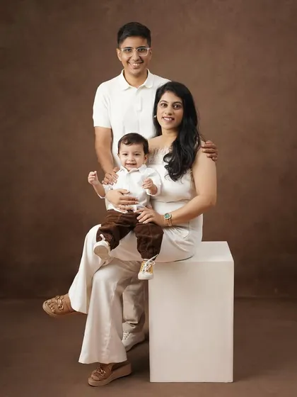 A formal family portrait against a solid brown backdrop, with the mother seated on a white block holding her son, and the father standing beside them.