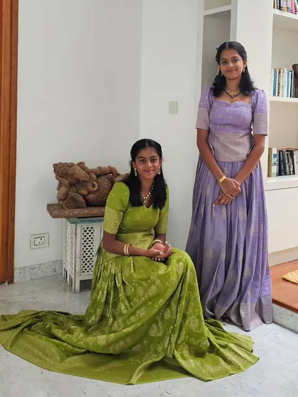 I design coordinated yet unique outfits for siblings. Here, two sisters wear custom lehengas in complementary shades of green and lavender. Each traditional lehenga is crafted from rich brocade fabric, reflecting their individual styles while maintaining a harmonious look.