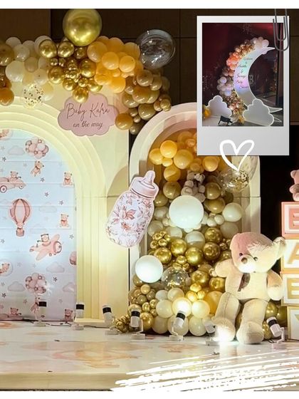 A close-up of the main stage, showing the custom-printed backdrop, a large baby bottle prop, and a plush teddy bear sitting among a sea of gold and white balloons.