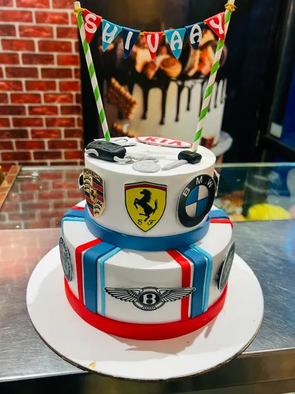 A two-tier cake for a luxury car enthusiast, decorated with the logos of Ferrari, BMW, Porsche, and Bentley.