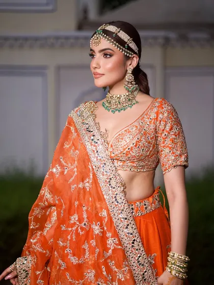 A portrait showcasing the beautiful contrast between the orange lehenga and the green emerald jewellery, a classic combination for Indian festivities.