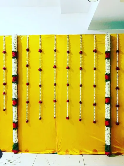 A minimalist yet traditional backdrop design. A bright yellow cloth is decorated with vertical garlands of tuberose and red roses.