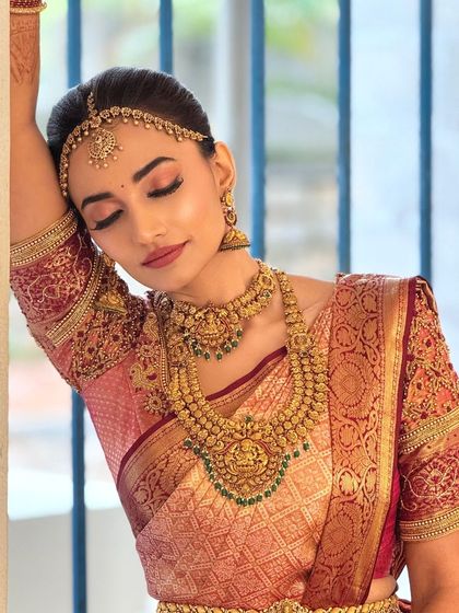 A serene portrait of a bride in a beautiful red and gold saree. The makeup features soft, rosy tones on the eyes and a defined lip.