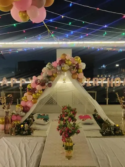 A birthday cabana on a rooftop decorated with a colorful balloon arch in pink and gold tones. The setup includes 'Happy Birthday' foil balloons and low seating for a relaxed and festive party.