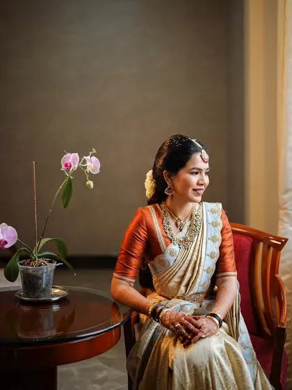 A beautiful, naturally lit portrait of a bride in a classic off-white and rust-orange silk saree. The makeup is kept soft and natural to enhance her features, creating a look of pure elegance.
