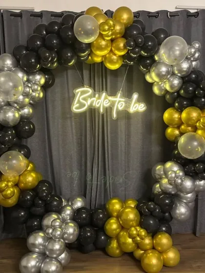 A chic and stylish 'Bride to be' ring backdrop for a bridal shower or bachelorette party. The circular frame is decorated with black, gold, and silver balloons and a central neon sign.