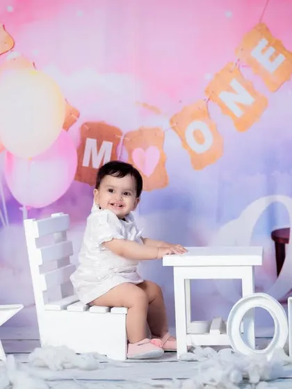 This baby girl is celebrating her first birthday with a "Mr & Mrs ONE" banner in the background, sitting at a tiny white table.