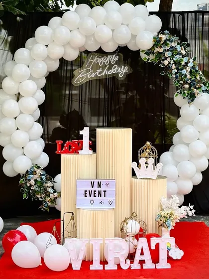 A beautiful outdoor birthday setup with a full white balloon arch accented with flowers. The black backdrop makes the white balloons and neon 'Happy Birthday' sign stand out.