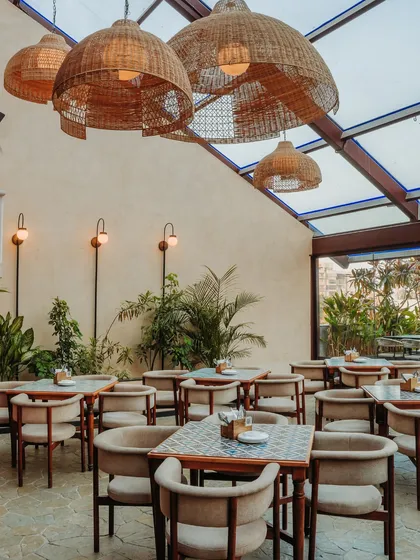 Under a soaring glass roof, this dining area is filled with natural light. The oversized woven pendant lamps create a warm, intimate atmosphere in the evening, while the patterned tabletops add a touch of eclectic charm.