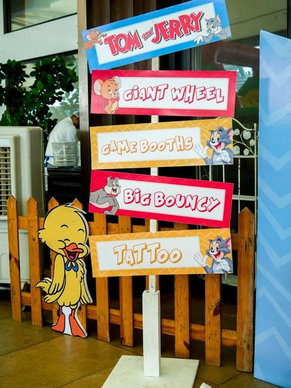 A Tom & Jerry themed signpost guiding guests to the "Giant Wheel," "Game Booths," "Big Bouncy," and "Tattoo" stations.