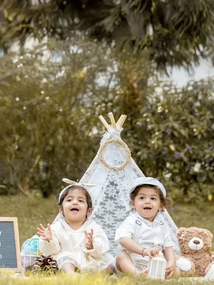 A happy, wide-angle shot of the twins enjoying their first birthday setup. Both are looking at the camera with big smiles, surrounded by all the fun props.