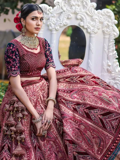 This portrait captures the bride seated in a garden, with a vintage mirror reflecting the surroundings. The composition is creative and highlights the rich texture and color of the velvet lehenga.