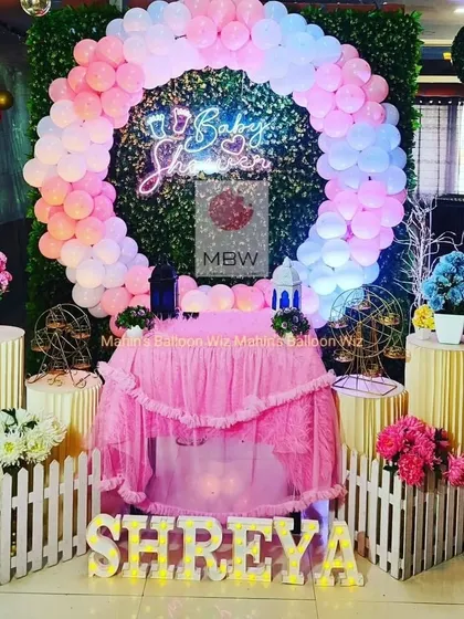 A classic baby shower cradle ceremony setup. The decor features a pink and blue balloon ring against a greenery wall, a neon "Baby Shower" sign, and a beautifully decorated cradle.