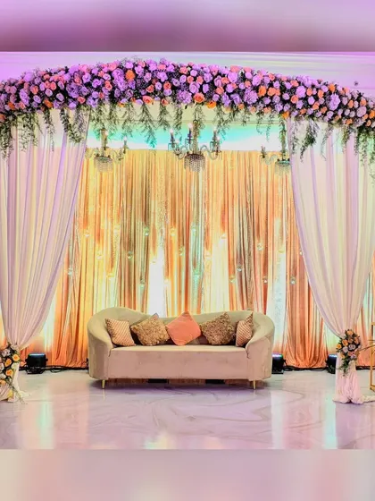 An elegant reception stage with a backdrop of soft, draped fabric, lit by warm uplighting and topped with a beautiful arrangement of peach and white flowers.