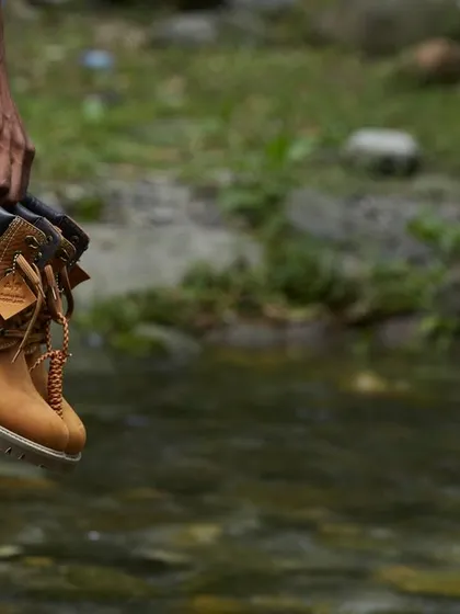 A product-focused shot for the Woodland "A Skating Monk" campaign. A hand holds the rugged leather boot against a natural, out-of-focus background of a stream, emphasizing the brand's adventurous spirit.