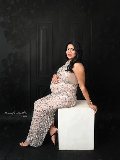 A chic and moody portrait. The mom-to-be is seated elegantly in a glamorous gown against a dark, textured backdrop, creating a sophisticated and modern look.