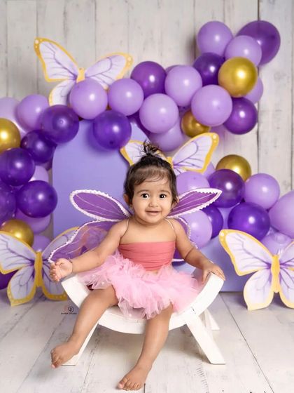 A sweet toddler dressed as a butterfly, smiling brightly for her milestone photoshoot. The purple and gold balloon arch and butterfly cutouts create a magical, whimsical setting.