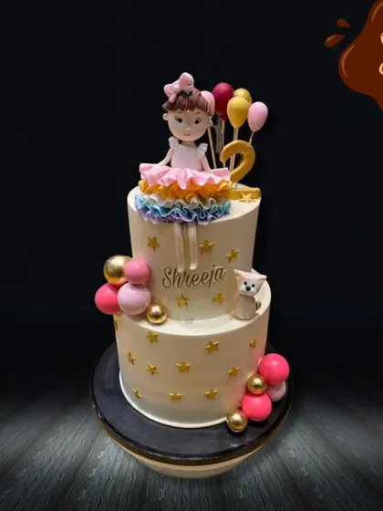 This elegant two-tier cake was designed for a sweet second birthday. It features a delicate doll topper, a cascade of rainbow ruffles, and balloon accents, all personalized with the birthday girl's name.