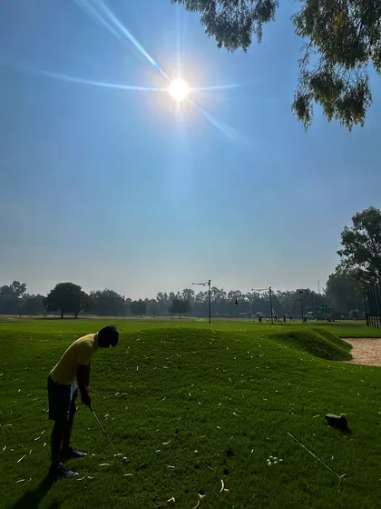 A young golfer practices his short game under the bright sun, with the bunker visible in the background.