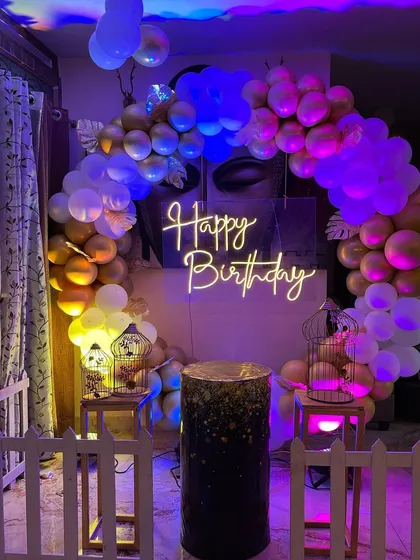 A circular balloon arch with a neon sign creates a fantastic focal point in this room. The setup is enhanced with purple and blue lighting, decorative birdcages, and a small picket fence for a whimsical touch.