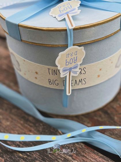 A close-up of the powder-blue hat box, showing the custom "Tiny Roars Big Dreams" band and "It's a boy" tags, all tied together with layered ribbons.
