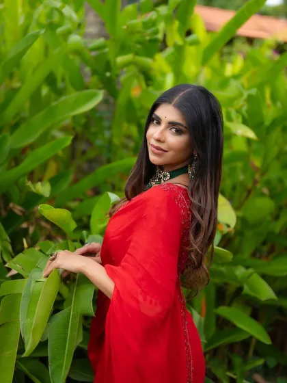 A beautiful portrait of an actress in a striking red saree. The natural green background makes the red color and the intricate jewelry details stand out.