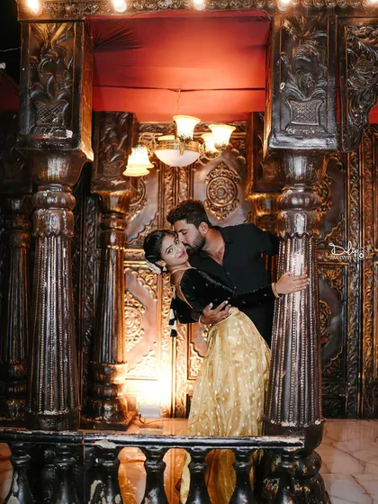 A romantic and glamorous shot on a balcony-like set, with the couple framed by ornate pillars and warm lighting.
