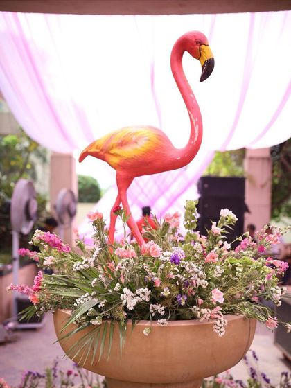 A playful and tropical floral centerpiece. A bright pink flamingo prop stands amidst a lush arrangement of pink and white flowers, adding a fun and quirky element to this poolside party decor.
