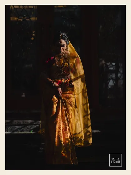 An artistic bridal portrait using light and shadow to create a dramatic effect. The bride, in her golden silk saree, is captured in a moment of quiet contemplation, highlighting her timeless elegance.