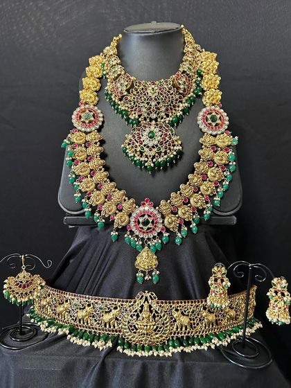 This is a very grand bridal set with multiple layers and intricate work. It features a choker, a mid-length necklace, and a long haaram, all adorned with floral motifs and green and red stones.
