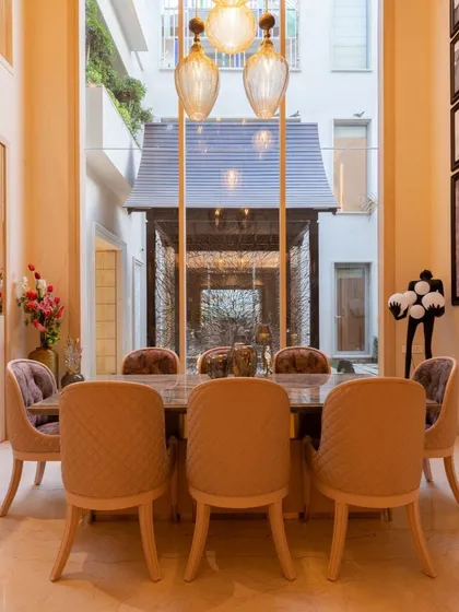 A double-height dining area that exudes elegance. The space is defined by a large window overlooking a courtyard, plush quilted chairs, and delicate pendant lighting, creating a grand yet intimate atmosphere.