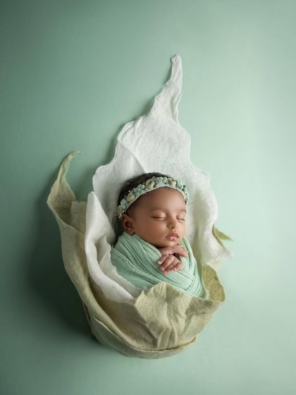 A newborn girl wrapped in a soft mint green swaddle, nestled inside a white and green flower prop. This creates a fresh, organic, and ethereal portrait.