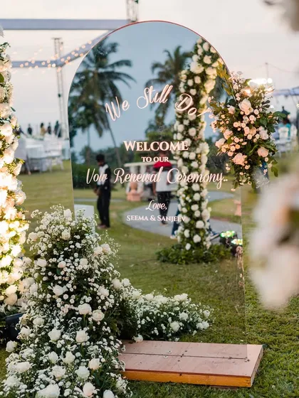 A simple and elegant welcome to a vow renewal ceremony. This custom acrylic sign, framed by beautiful white floral arrangements, sets a romantic and personal tone for a "We Still Do" celebration against a Goa sunset.