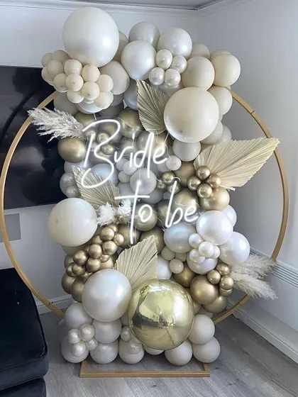 A chic and modern "Bride to be" setup. The gold ring is decorated with an arrangement of white and gold balloons and dried palm leaves, with a neon sign as the centerpiece.
