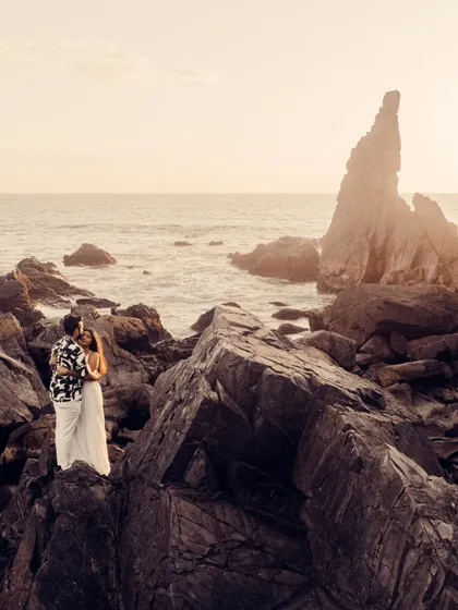 A wide shot of a couple standing on the dramatic rock formations of a Goa beach at sunset. This image showcases the epic scale and beauty of a destination pre-wedding shoot.