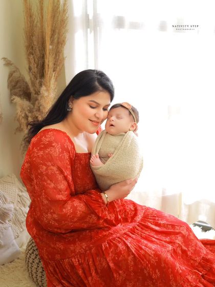 A mother in a beautiful red dress holds her sleeping baby. The contrast of the vibrant dress against the neutral, bohemian-style background makes for a striking portrait.