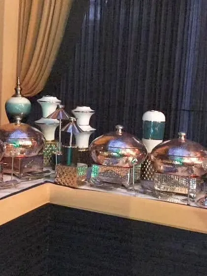 A corner view of an elegant buffet table with polished copper chafing dishes and decorative ceramic vases, set against a backdrop of dark curtains.