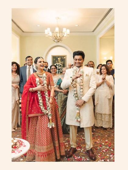 A joyful moment after the Varmala exchange. The couple stands together, officially united, surrounded by the happy faces of their family and friends in an elegant indoor setting.