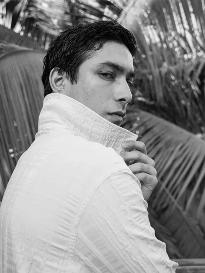 A classic black and white male portrait, shot outdoors against natural foliage. The over-the-shoulder gaze is engaging, and the popped collar adds a touch of style.