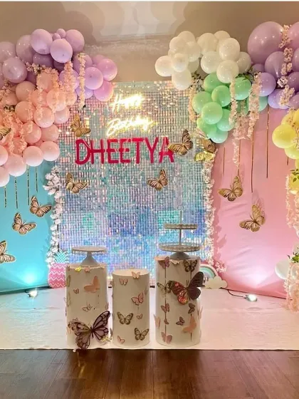 A vibrant butterfly-themed party featuring a stunning iridescent shimmer wall that catches the light beautifully. The setup is framed by a pastel rainbow balloon garland and includes themed plinths for the cake.