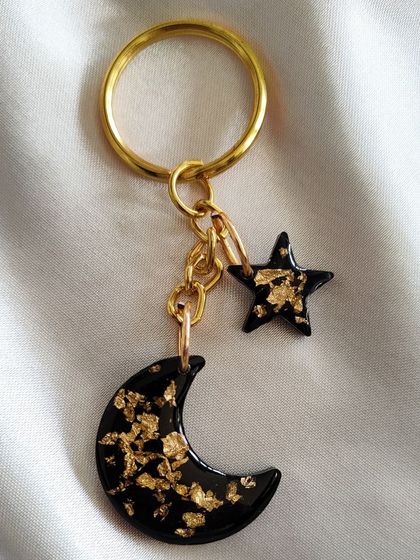 A close-up of the black and gold moon and star keychain. The gold flakes stand out beautifully against the dark, glossy background.