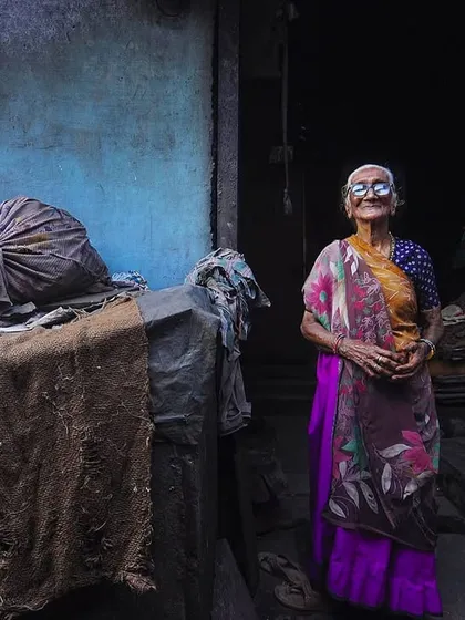 An elderly woman stands in a dimly lit alley, her gentle smile illuminated by a sliver of light. Her portrait tells a story of a long life lived with grace and strength.