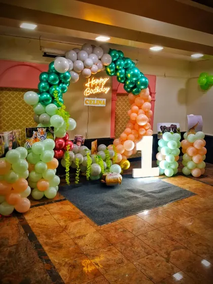 This jungle-themed decoration for a first birthday uses a soft, earthy color palette of green, orange, and white balloons. The arrangement includes a large light-up number '1' and is designed to create a fun, safari-like atmosphere for the little one's big day.